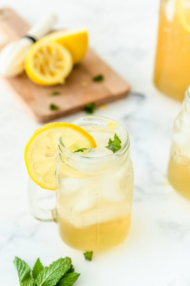vodka lemonade in a glass mug garnished with lemon. squeezed lemons and fresh mint in background