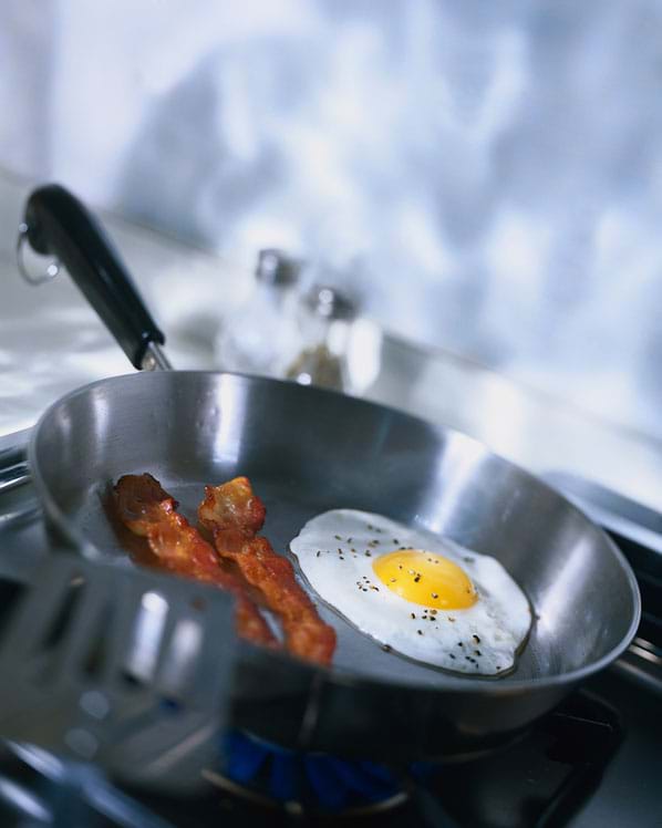 Photo shows bacon and eggs being cooked in a saucepan on a stove burner.